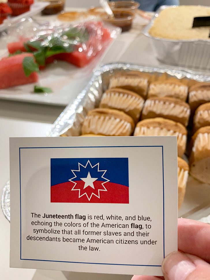 Close-up of a table of delicious food with a hand holding an informative card that reads, "The Juneteenth flag is red, white, and blue, echoing the colors of the American flag, to symbolize that all former slaves and their descendants became American citizens under the law."