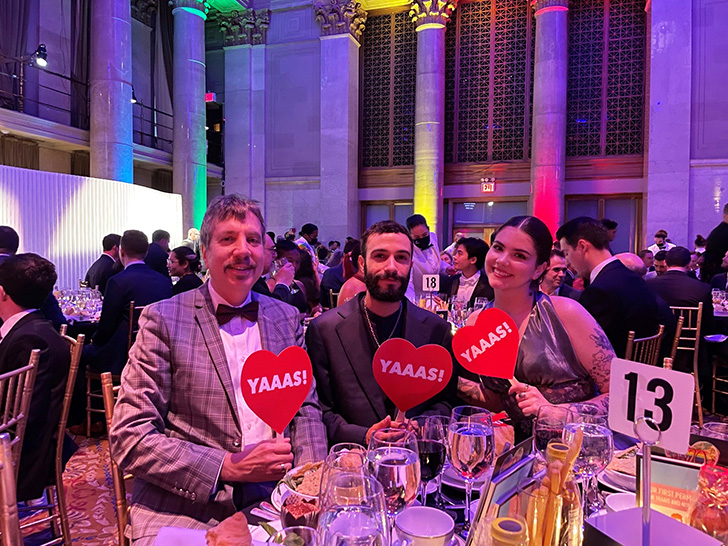 Three staff members from our Chelsea store hold heart-shaped "Yaaas!" signs at a table during a dinner gala.